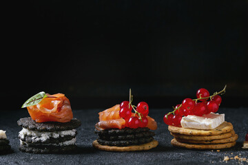 Stack of black wholegrain charcoal and traditional crackers with smoked salmon, cream cheese, green salad and red currant berries over black stone background. Appetizer snack. Space for text