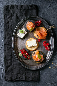 Stack of black charcoal and traditional crackers with smoked salmon, cream cheese, green salad and red currant berries on vintage metal tray over black stone background. Appetizer snack. Top view