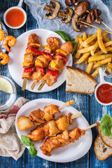 Variety of BBQ snack lunch. Plates grilled spicy prawn kebabs, chicken, pork, vegetables, mushrooms skewers, bread, french fries potatoes with sauces and greens over blue wooden background. Flat lay