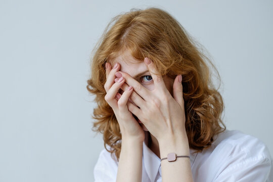 A red-haired girl with curls in embarrassment covers her face with her hands and looks through her fingers. Social phobia of the younger generation.
