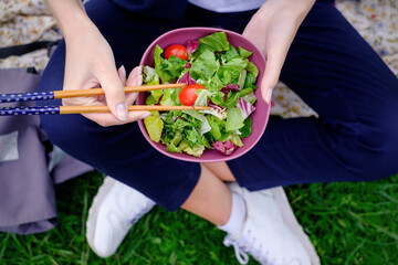 A girl sits on the grass and eats a green fresh salad with chopsticks for sushi. A healthy diet on plant foods.