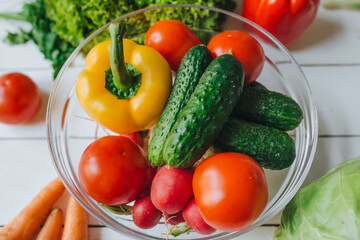 Fresh vegetables in glass bowl on white wooden table top view. red, yellow, green colors. Vegan concept.