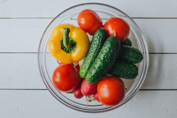 Fresh vegetables in glass bowl on white wooden table top view. red, yellow, green colors. Vegan concept.