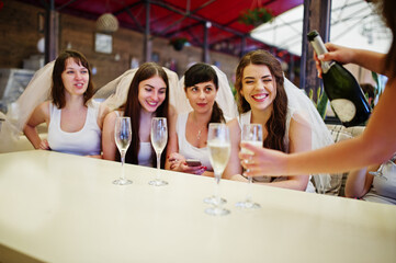 Group of cheerful girls at white shirts sitting at table and drink champagne on hen party.