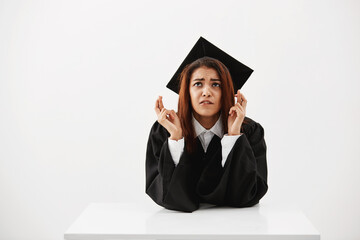 Nervous african student hoping to get her diploma, with fingers crossed over white wall sitting at table. Education concept.