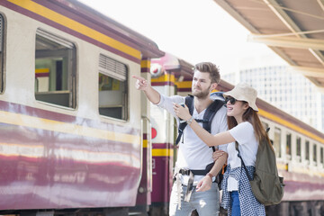 Fototapeta na wymiar Young couple asian woman and caucasian man traveler are pointing and looking out with train station background. Travel in summer concept. Lover concept.