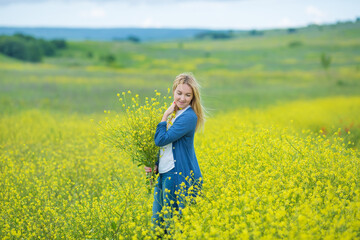 Naklejka na ściany i meble Genuine cute lady woman in meadow of yellow flowers sniffing flower bouquet. Attractive beautiful young girl enjoying the warm summer sun in a wide green and yellow meadow. Stylish dressed