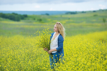 Fototapeta premium Genuine cute lady woman in meadow of yellow flowers sniffing flower bouquet. Attractive beautiful young girl enjoying the warm summer sun in a wide green and yellow meadow. Stylish dressed