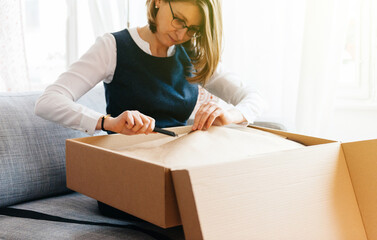 Young beautiful woman on living room sofa using cutter as she unpacks unbox cardboard box parcel...