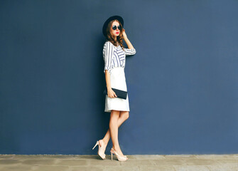 Fashion elegant woman in a black hat and white skirt with a handbag clutch over a gray background