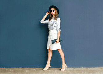 Fashion elegant woman wearing a black hat and white skirt with a handbag clutch walking over a gray background