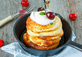 Homemade cottage cheese pancakes with sour cream and cherries in a cast-iron frying pan. Russian dessert from cottage cheese.