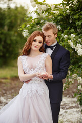 Young Bride and Groom couple in a blooming garden. Tender holding each other. Spring wedding. Redhead girl with long hairs. Young family outdoor image near blooming bush of spirea. Love and tenderness