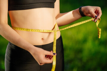 Young woman measuring her thin waist with a tape measure on green nature backgroung, close up. Diet...