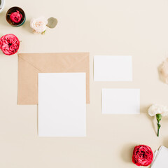 Wedding invitation cards, craft envelope, pink and red rose flower buds and white carnation on pale pastel beige background. Workspace with paper blank. Flat lay, top view
