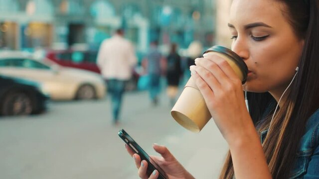 Beautiful young woman listening yo music on smart phone, moving to the rythm and drinking coffe while sitting on the steps outside on the street. Close up