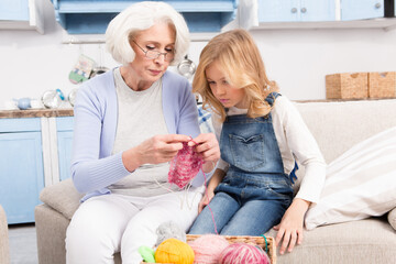 Closeup of grandmother showing her granddaughter how to knit jumper or vest while sitting on sofa or couch in living room at home.