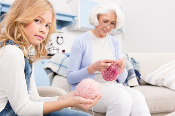 Obraz premium Granddaughter holding ball of threads and helping her grandmother. Granny knitting different kinds of clothes for whole family.