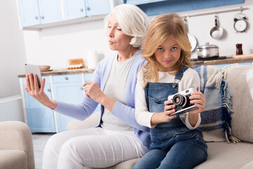 Grandmother and granddaughter studies computer gadgets of each other. Pretty ladies taking pictures on different gadgets.