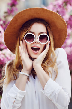 Outdoor portrait of young beautiful girl looking surprised, shocked. Model expressing joy, excitement. Lady wearing stylish round sunglasses, hat, posing in street, near blooming tree. City lifestyle