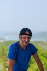 Naklejka premium Happy Young Bearded Man standing on view point with a seascape wearing in blank blue t-shirt and black Cap