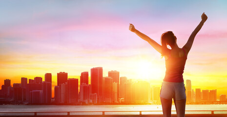 Fototapeta na wymiar Jogging, Fitness And Success - Girl With Arms Raised And Cityscape At Sunset