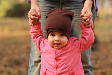 Portrait of a cute baby girl holding mom's hands and learning to walk in the spring park
