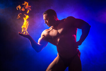 A man with an inflated torso. Handsome bodybuilder with fire in hands. Bodybuilder on a blue background. - obrazy, fototapety, plakaty