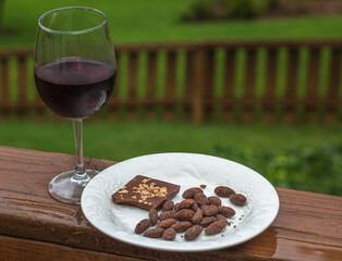 Chocolate covered almonds and chocolate almonds on a white plate with a glass of red wine on a wooden porch after a spring rain