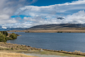 Middle Earth, New Zealand - March 14, 2017: Wide  blue Lake Clearwater in front. Volcano Potts peaking through the white clouds in a blue sky in the back. Green and beige vegetation.