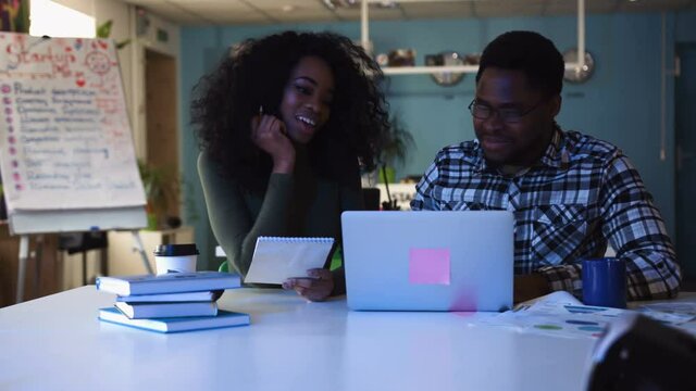 A young African American man with glasses and beautiful young African American woman with curly hair talking about business plans and working on a laptop in the office at night smiling