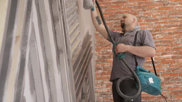 Young European makes repairs in apartment washing crossbeams. Man with beard, dressed in typical clothes for repairs: gray T-shirt and black trousers, cleans metal shelves on the wall with spray gun