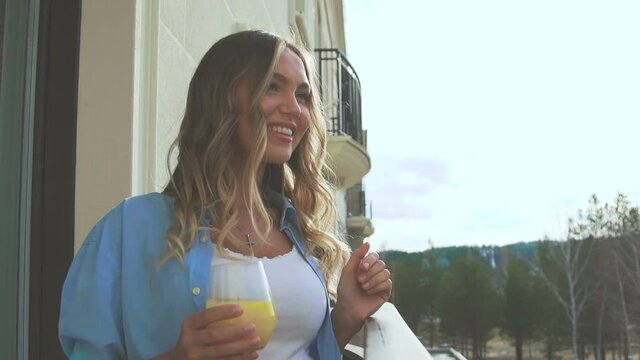 Woman on the balcony in the early morning and drinks orange juice. Attractive blonde drinking fresh on terrace. A romantic girl is standing on the veranda, looking into the distance and drinking juice