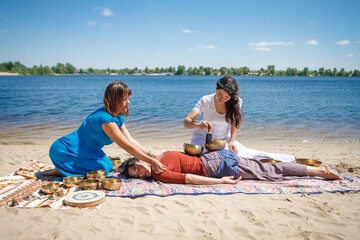 Obraz premium Beautiful female receiving energy sound massage with singing bowls and body massage on a river bank at spring sunny day