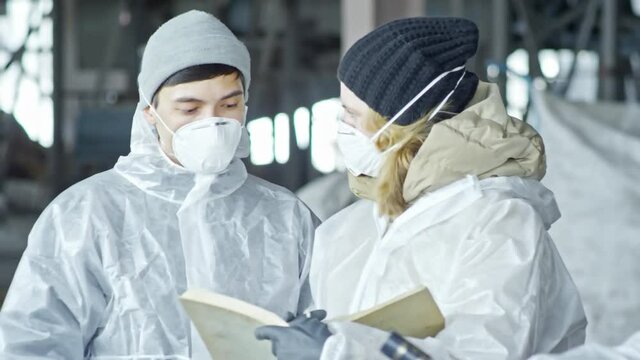 Medium shot with PAN of woman and man in protective coveralls and masks discussing work and making notes as other workers sorting waste at recycling facility 