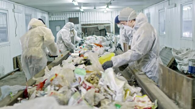 PAN of people in protective disposable coveralls standing before moving conveyor belt full of waste and sorting plastic 
