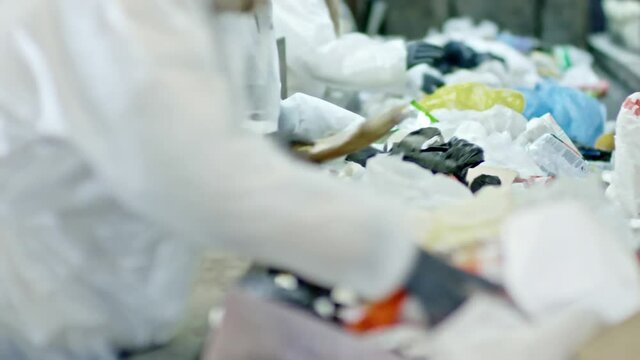 PAN with slow motion of unrecognizable people in protective coveralls and masks working at moving conveyor belt with piles of household waste and sorting plastic bottles 