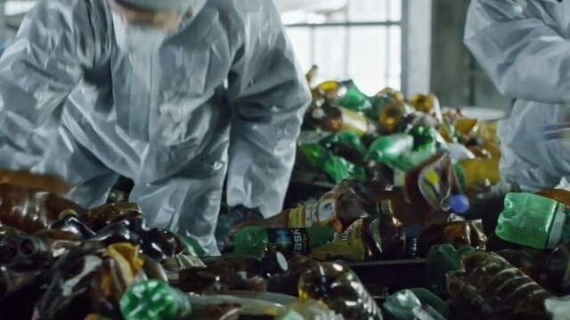 Slow motion shot with PAN of unrecognizable garbage recycling facility workers in protective coveralls and mask standing in container full of plastic bottles and throwing them out
