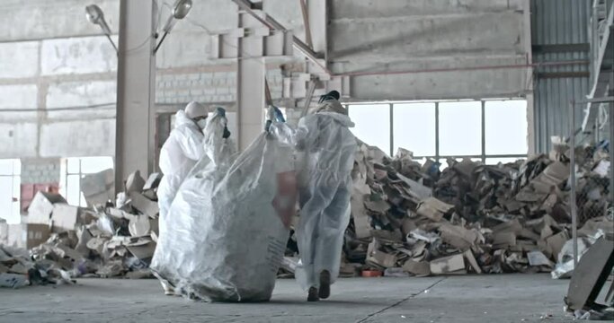 Slow motion dolly shot of garbage recycling factory workers in protective coveralls and masks carrying bag of litter towards cardboard waste as their colleague sorting plastic litter 