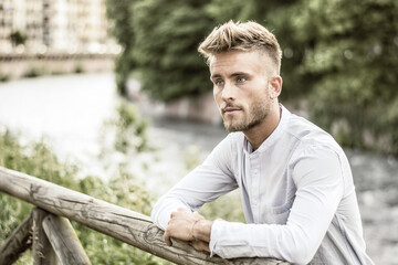 Fototapeta premium Blondish, blue eyed young man by river at sunset, thinking, looking away