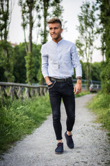 Handsome young man in white shirt and black pants outdoor in city park, walking toward camera, serious