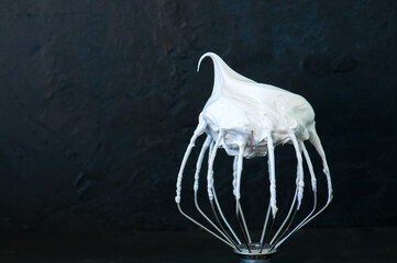 Whipped egg whites - beaten italian merengue on a whisk on black table,  closeup and copy space.