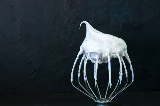Whipped egg whites - beaten italian merengue on a whisk on black table,  closeup and copy space.