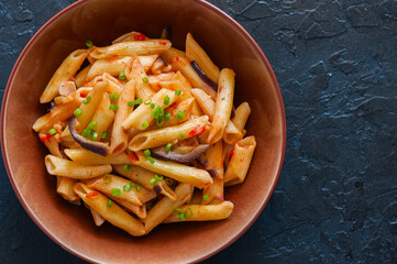 Close up of italian penne pasta with eggplants and tomato sauce in a brown bowl on a black stone background. Copy space and overhead view. Traditional food concept.