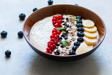 Healthy oat smoothie with bananas and cottage cheese garnished with red currants, sunflower seeds, chocolate drops and blueberries in a bowl on a white stone background.