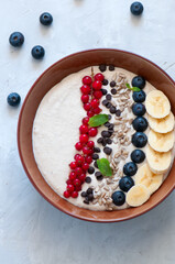 Healthy oat smoothie with bananas and cottage cheese garnished with red currants, sunflower seeds, chocolate drops and blueberries in a bowl on a white stone background.