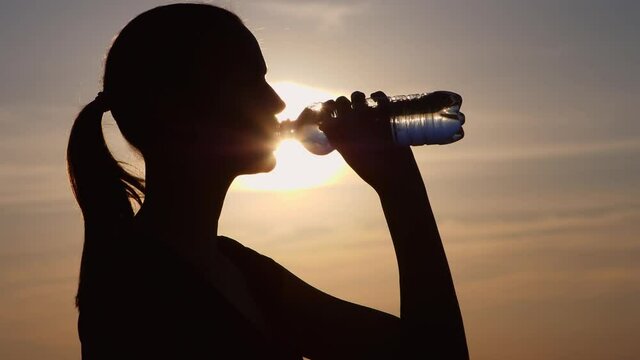 Fitness woman drinking water outdoors at sunset. Silhouette of a girl drinking water from a bottle. Slow motion