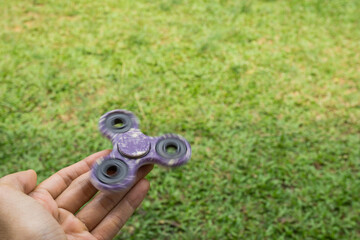A hand holding fidget spinner purple colour spinning stress relieving toy on green grass background.