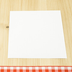 Ein weißer Zettel auf Holzhintergrund, am unteren Bildrand ein rot weiß kariertes Band, quadratisch, Textfreiraum