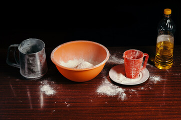 Orange cup, measuring cup, and a steel sieve, sunflower oil stand on a wooden table on a black background. Flour.